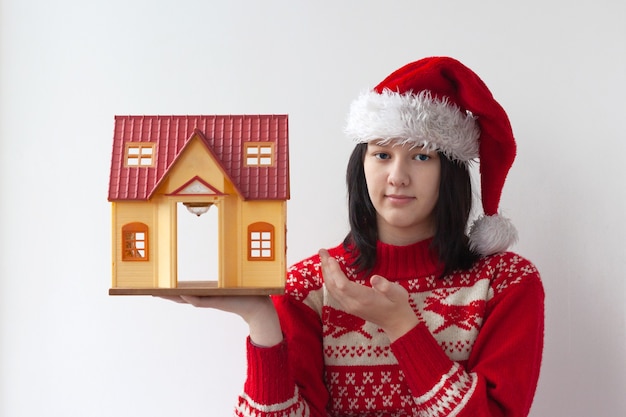 Une adolescente dans un chapeau de Père Noël et un pull de Noël rouge tient une maison de jouets. Le concept de Noël et du nouvel an