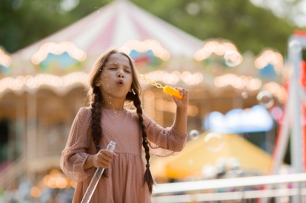 Adolescente avec deux nattes lance des bulles dans un parc d'attractions près de carrousel et se réjouit