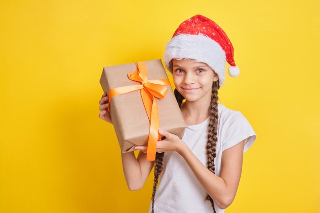 Adolescente émotionnelle en chapeau de père noël avec un cadeau sur fond jaune, concept de cadeaux de noël