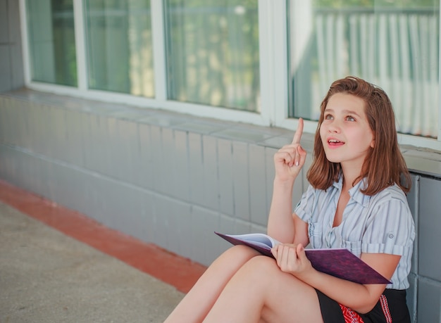 Photo une adolescente est assise à la fenêtre de l'école et lit un livre. l'enfant leva le doigt. écolière est venu à l'esprit une bonne idée. l'enfant a compris la leçon et a fait ses devoirs.