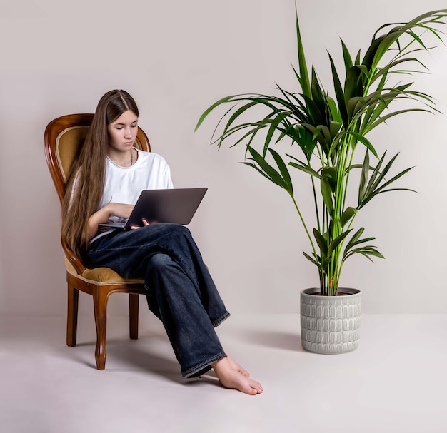 Une adolescente est assise à la maison dans un fauteuil avec un ordinateur portable en jeans et un t-shirt blanc, discutant pieds nus ou étudiant en ligne via Internet