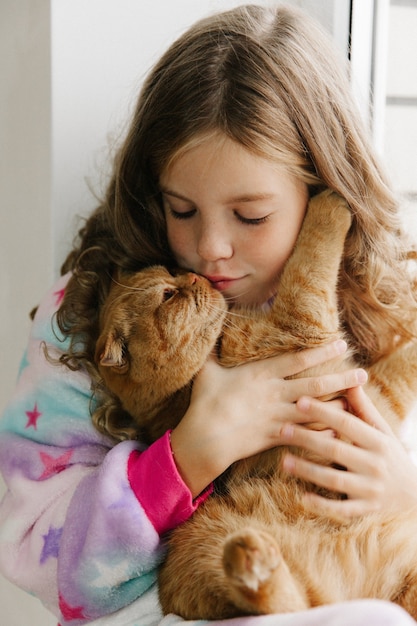 Une adolescente est assise en pyjama à la fenêtre de la maison et tient un chat roux. rester à la maison.