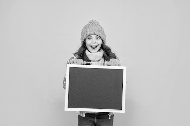 Une adolescente étonnée en vêtements d'hiver tient le tableau noir de l'école avec de la publicité pour l'espace de copie