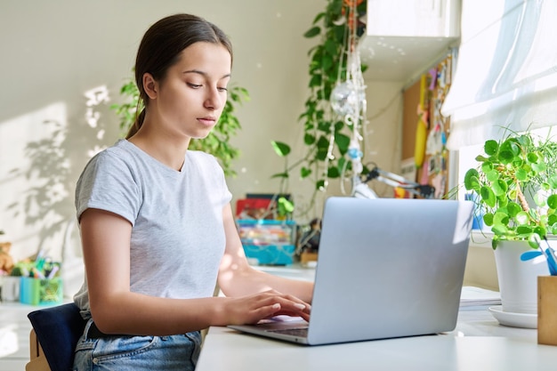 Adolescente étudiant à la maison à table à l'aide d'un ordinateur portable Étudiante adolescente étudiant en ligne faisant ses devoirs en écrivant dans un cahier en regardant une leçon d'écoute vidéo Elearning adolescence technologie du lycée