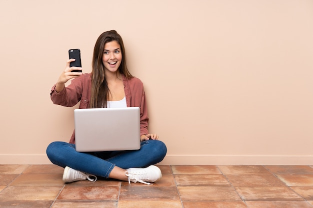 Adolescente étudiante assise sur le sol avec un ordinateur portable faisant un selfie