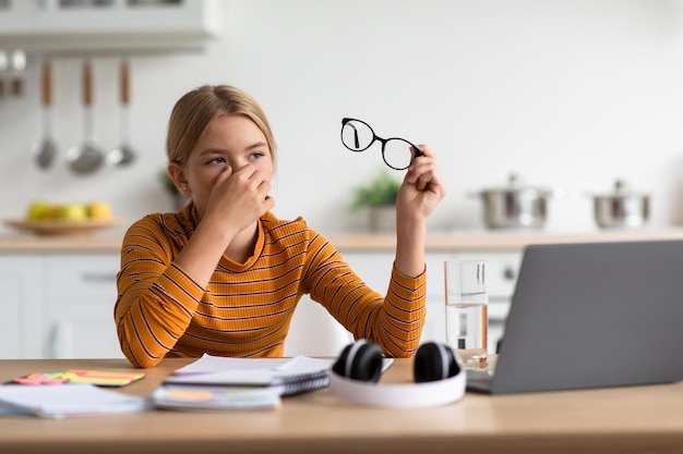 Une adolescente européenne fatiguée blonde enlève ses lunettes et se frotte les yeux à table avec un ordinateur portable dans la cuisine