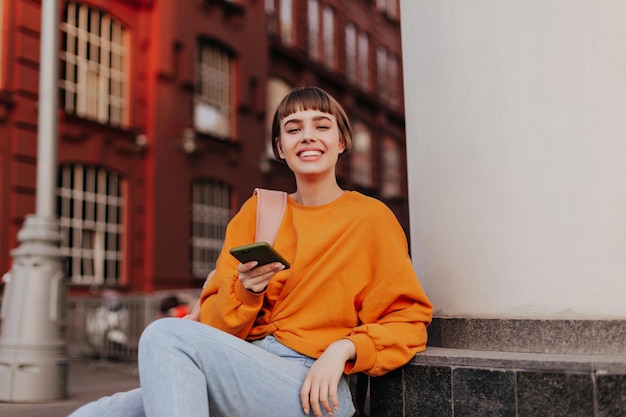 Une adolescente excitée en sweat-shirt orange s'appuie sur la construction et tient un smartphone Joyeuse femme en tenue hipster élégante sourit à l'extérieur