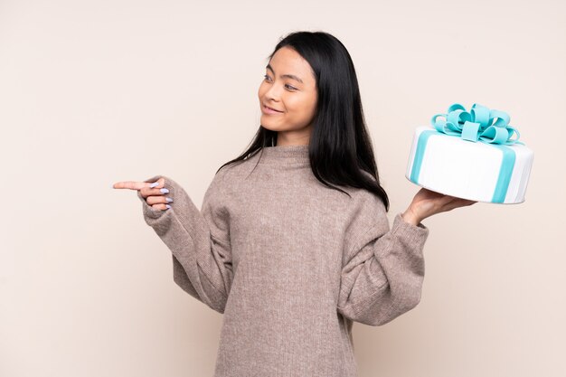 Adolescente fille asiatique tenant un gros gâteau sur un mur beige pointant vers le côté pour présenter un produit