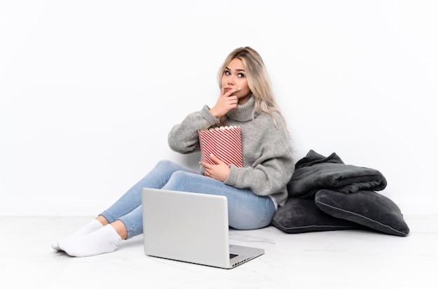 Adolescente fille blonde mangeant du pop-corn tout en regardant un film sur l'ordinateur portable ayant des doutes et avec une expression de visage confuse
