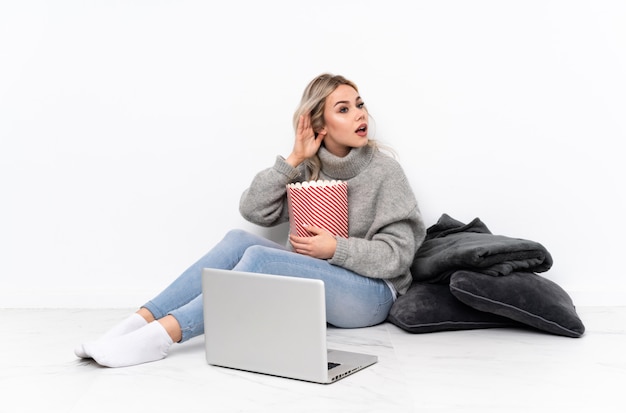 Adolescente fille blonde mangeant du pop-corn tout en regardant un film sur l'ordinateur portable en écoutant quelque chose en mettant la main sur l'oreille