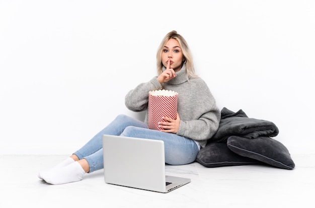 Adolescente fille blonde mangeant du pop-corn tout en regardant un film sur l'ordinateur portable montrant un signe de silence mettant le doigt dans la bouche