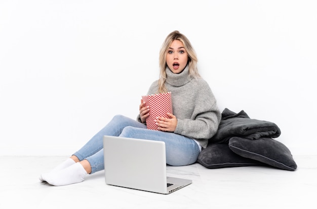 Adolescente fille blonde manger du pop-corn tout en regardant un film sur l'ordinateur portable avec une expression faciale surprise