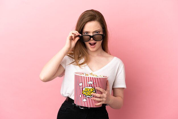 Adolescente sur fond rose isolé surpris avec des lunettes 3d et tenant un grand seau de pop-corn