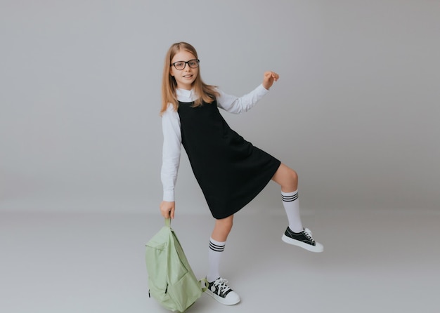 Adolescente gaie de 111213 ans dans un uniforme scolaire porte un sac à dos sur un fond gris mode scolaire