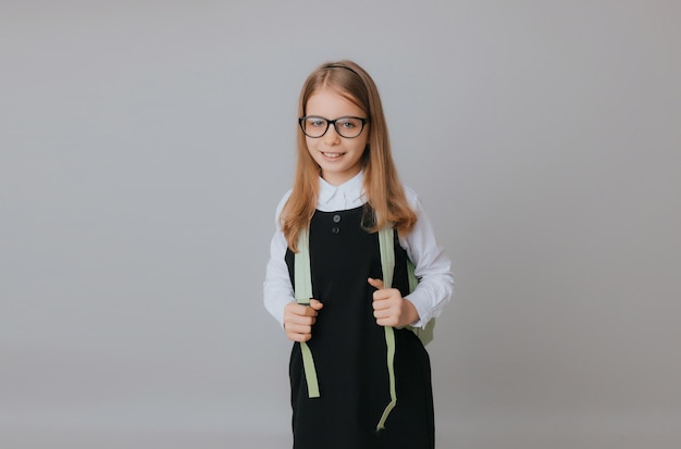 Adolescente gaie de 111213 ans dans un uniforme scolaire porte un sac à dos sur un fond gris mode scolaire