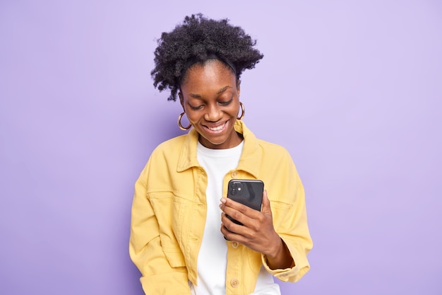 Une adolescente gaie et bouclée utilise un téléphone portable pour faire des achats en ligne ou envoyer des SMS sourit joyeusement a une expression heureuse