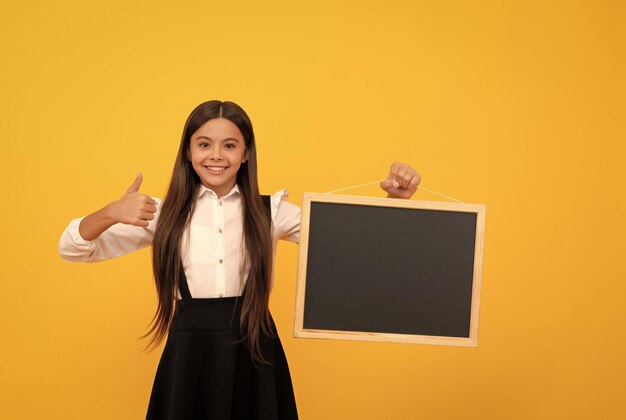 Adolescente gaie en uniforme tenir le tableau noir de l'école pour l'espace de copie montrant le pouce vers le haut présentant