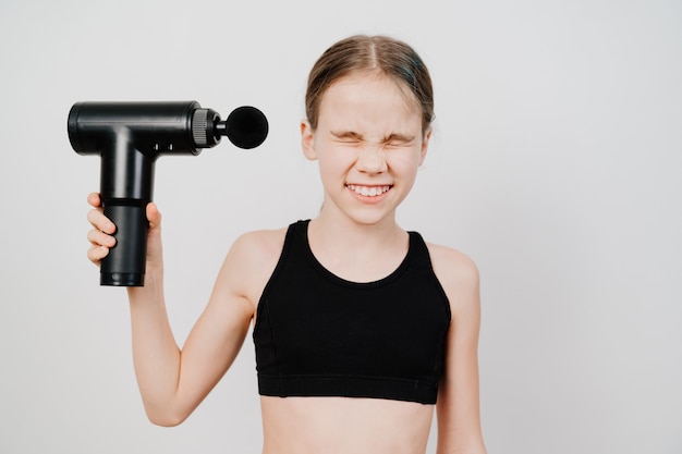 Une adolescente grimace et tient un pistolet de massage. appareil médico-sportif aide à réduire les douleurs musculaires après l'entraînement, aide à soulager la fatigue, affecte les zones à problèmes du corps, améliore l'état de la peau.