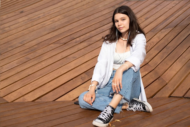 Une adolescente en gros plan dans des vêtements décontractés et des baskets écoute de la musique avec des écouteurs et s'assoit sur un banc en bois en plein air