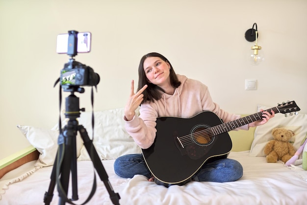 Adolescente avec guitare acoustique assise à la maison sur le lit, enregistrant de la musique en vidéo et discutant avec des amis