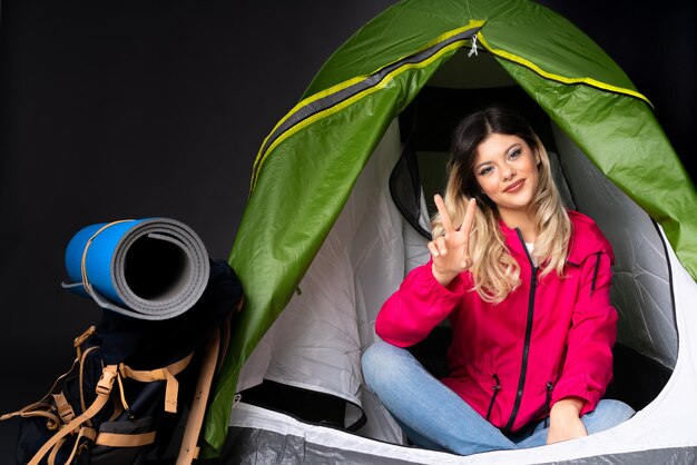 Adolescente à l'intérieur d'une tente de camping vert sur mur noir souriant et montrant le signe de la victoire