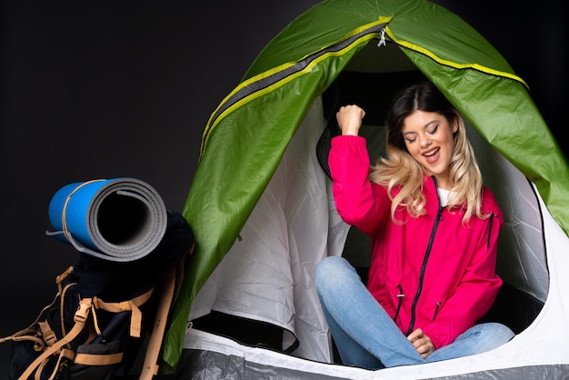 Adolescente à l'intérieur d'une tente de camping verte isolée sur un mur noir célébrant une victoire