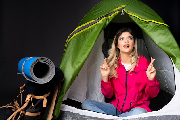 Adolescente à l'intérieur d'une tente de camping verte sur un mur noir avec les doigts se croisant et souhaitant le meilleur