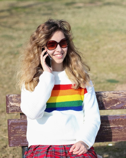 Photo une adolescente jolie et souriante qui parle au téléphone portable une jeune fille joyeuse et bouclée qui porte un pull arc-en-ciel