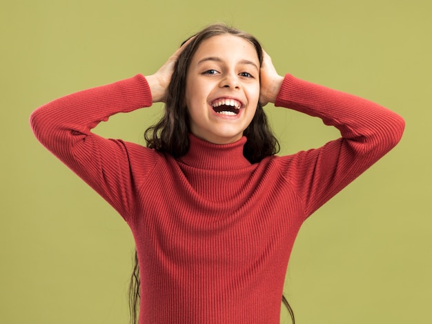 Adolescente joyeuse gardant les mains sur la tête regardant l'avant en riant isolé sur un mur vert olive