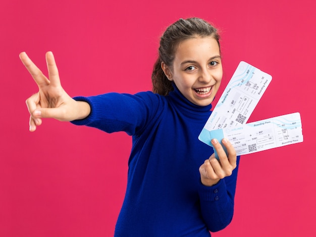 Photo adolescente joyeuse tenant des billets d'avion regardant à l'avant faisant un signe de paix isolé sur un mur rose
