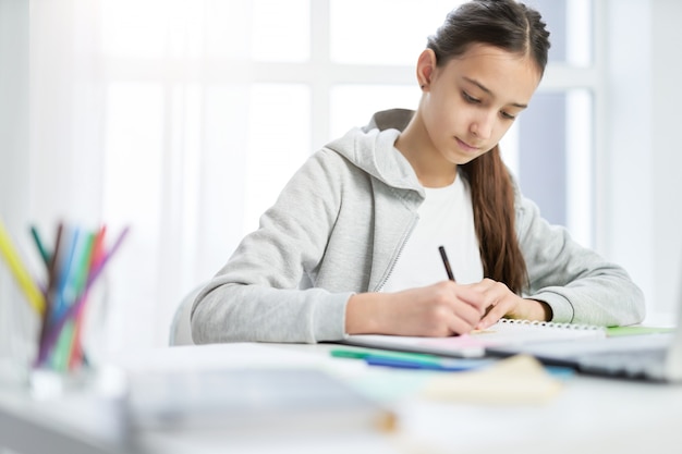 Adolescente latine concentrée prenant des notes dans son cahier tout en faisant ses devoirs, assise à la table à la maison. Enseignement à distance, concept d'enseignement à domicile