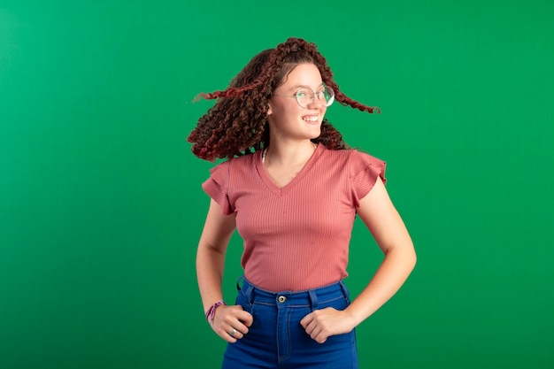 Adolescente avec des lunettes berçant ses cheveux roux bouclés dans des poses amusantes en studio tourné avec un fond vert idéal pour le recadrage