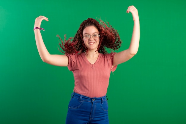 Adolescente avec des lunettes berçant ses cheveux roux bouclés dans des poses amusantes en studio tourné avec un fond vert idéal pour le recadrage