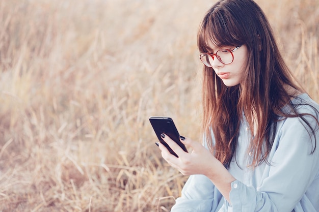 Une adolescente avec des lunettes est assise sur la pelouse et regarde le téléphone