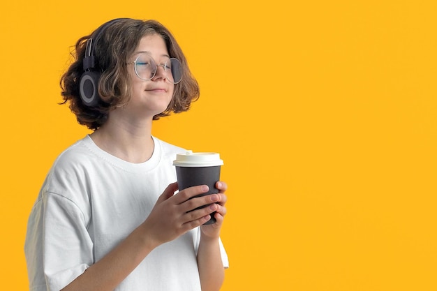 Une adolescente avec des lunettes tient une tasse de café dans sa main et écoute de la musique avec des écouteurs