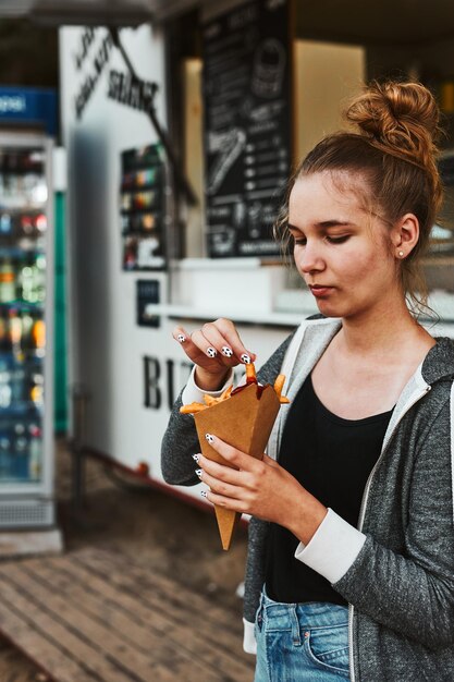 Photo une adolescente mangeant des pommes de terre frites à partir d'un cône en carton devant un camion de nourriture