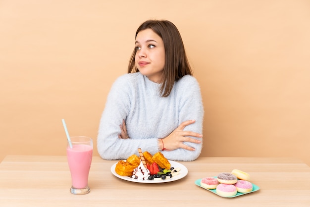 Adolescente, manger des gaufres sur un mur beige, faisant des doutes, tout en soulevant les épaules