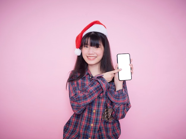Adolescente mignonne en bonnet de Noel tenant le smartphone avec écran blanc sur le mur rose. Espace pour le texte