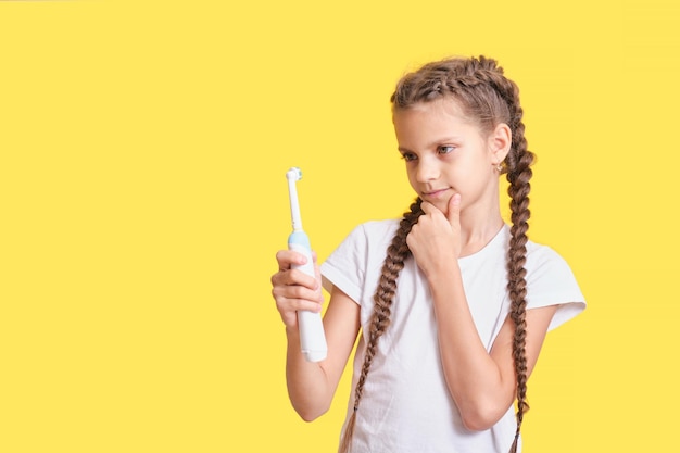 Adolescente mignonne avec la coiffure de nattes tenant une brosse à dents électrique dans ses mains sur un fond jaune, sélection de produits d'hygiène buccale pour les enfants