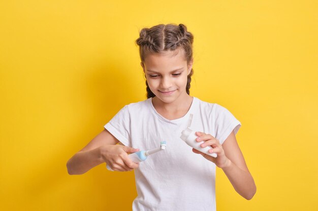 Une adolescente mignonne avec une coiffure en tresses serre le dentifrice sur une brosse à dents électrique, fond jaune, gros plan, mise au point sélective, hygiène buccale des adolescents