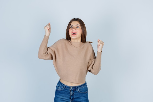 Adolescente montrant le geste du gagnant en pull, jeans et l'air heureux, vue de face.