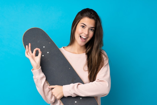 Adolescente sur mur bleu isolé avec un patin et faisant signe OK