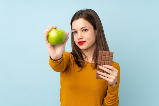 Adolescente sur mur bleu en prenant une tablette de chocolat dans une main et une pomme dans l'autre