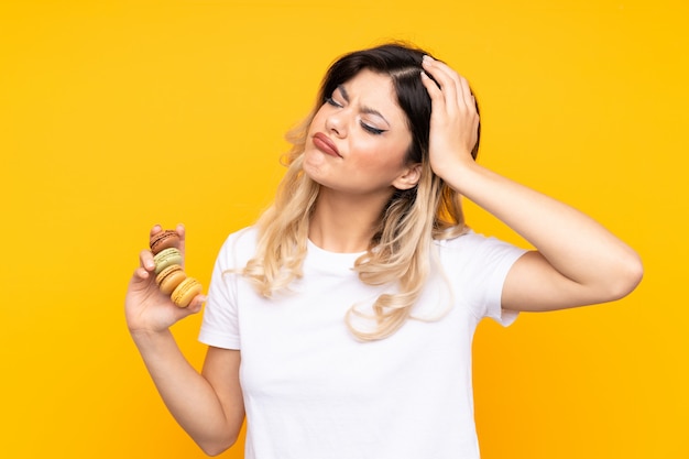 Adolescente sur mur jaune tenant des macarons français colorés avec une expression confuse