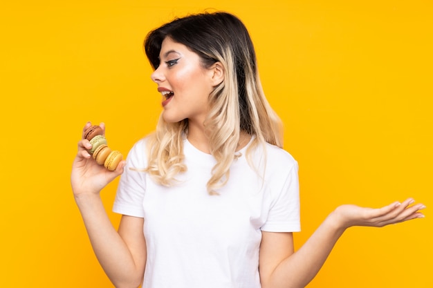 Adolescente sur mur jaune tenant des macarons français colorés et avec une expression surprise