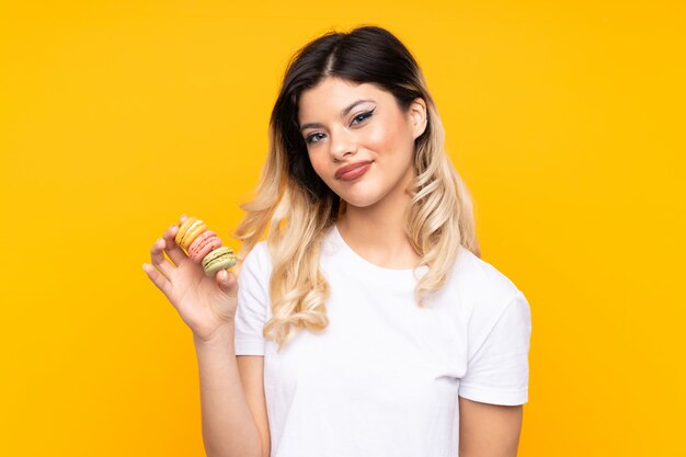 Adolescente sur mur jaune tenant des macarons français colorés avec une expression triste