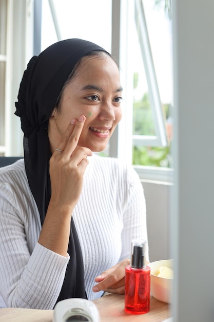 Adolescente musulmane joyeuse et souriante faisant une routine de soins de la peau, appliquez du sérum et de la crème sur le visage à la maison