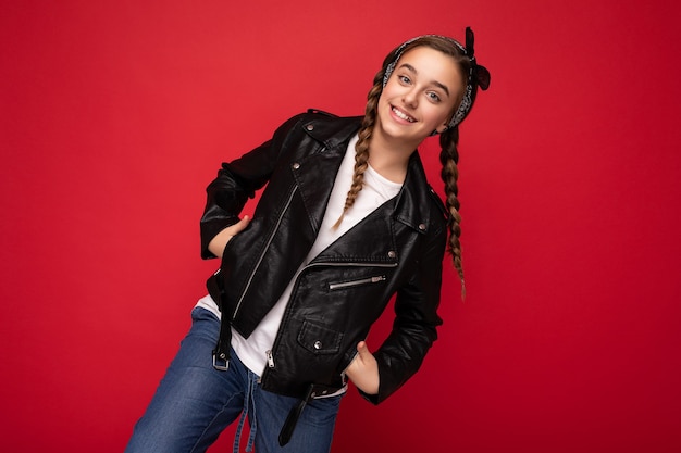 Adolescente avec des nattes portant élégante veste en cuir noir et tshirt blanc isolé sur fond rouge mur regardant la caméra.