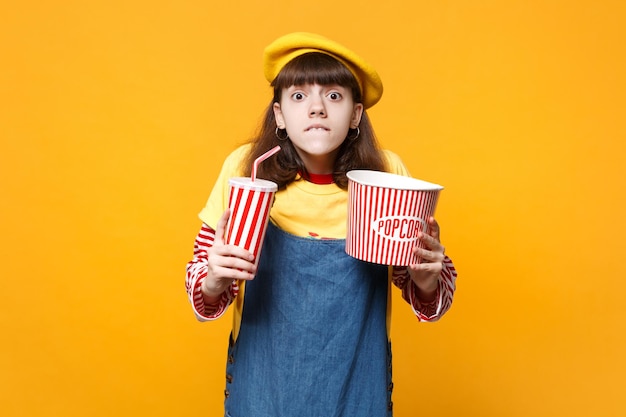 Adolescente nerveuse en béret français se mordant les lèvres, tenant une tasse en plastique de cola ou de soda, seau de pop-corn isolé sur fond jaune. Émotions sincères des gens, concept de style de vie. Maquette de l'espace de copie.