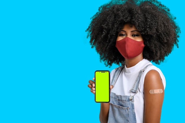 Adolescente noire tenant un téléphone portable à côté de son bras avec un autocollant de vaccin qu'elle porte une cellule de protection faciale